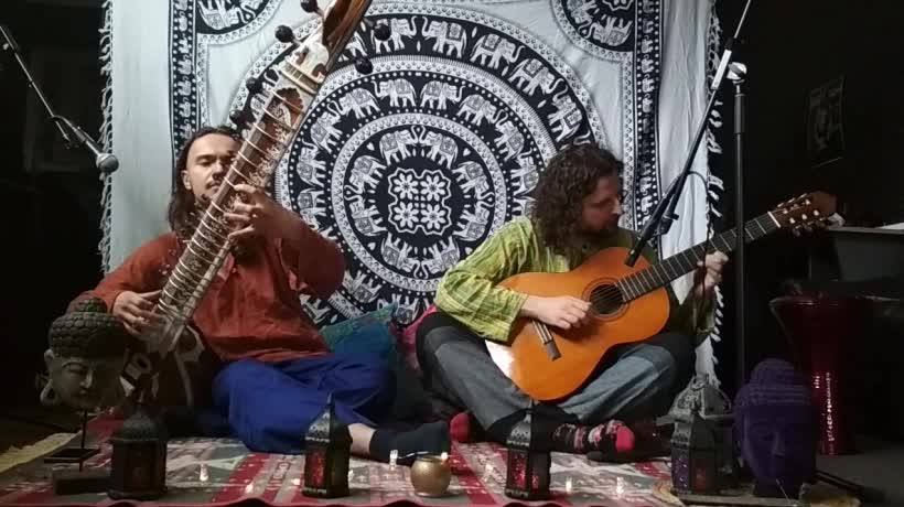 Doi muzicieni cântă la sitar și chitară la lumina lumânărilor, pe fundalul unei cuverturi indiene