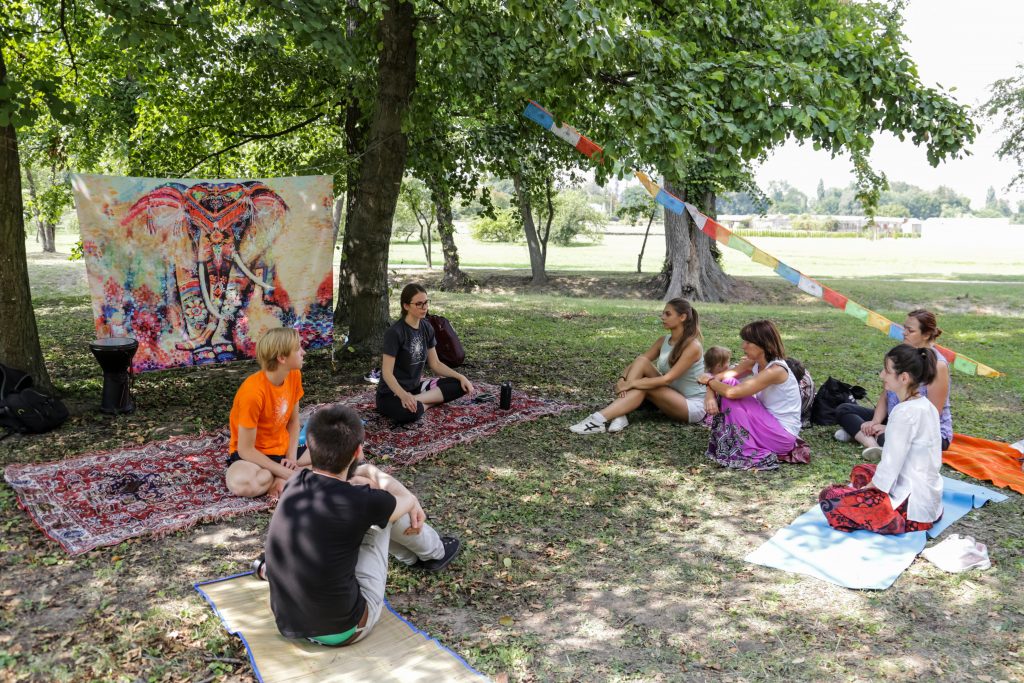 Un grup mic stă în cerc pe saltele în parc, lângă o pânză cu un elefant, și ascultă o prelegere