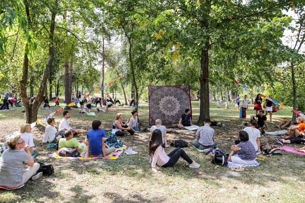 Participanții la festival stau pe saltele în parc, sub copaci cu ghirlande de stegulețe, iar în fundal se practică yoga