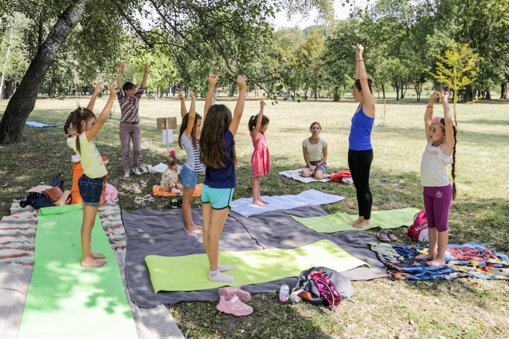 Copiii și instructoarea practică yoga pentru copii pe saltele, în aer liber într-un parc.