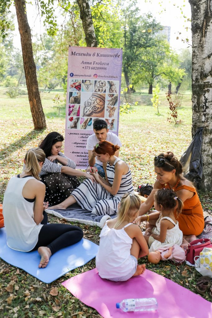 Femei și copii stau pe saltele în parc, în timp ce un artist aplică un desen cu mehendi pe mână.